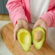 Mulher com um abacate cortado ao meio nas mãos em cima de uma mesa com verduras