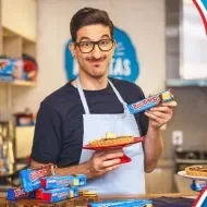Imagem de um rapaz alto com uma torta e um avental azul à esquerda e à direita o nome do programa em letras vermelhas