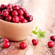 Tigela de madeira com cranberries frescos sobre superfície rústica, com algumas frutas espalhadas ao redor.