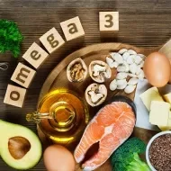 Alimentos ricos em ômega-3 sobre uma tábua: salmão, abacate, ovos, oleaginosas, azeite, manteiga e brócolis.