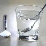 Copo com água e colher dentro, ao lado de colher com sal sobre uma mesa clara, representando soro caseiro.