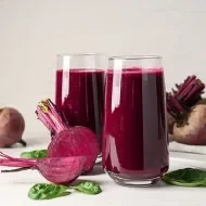 Copos com suco de beterraba, rodeados por beterrabas frescas cortadas e folhas verdes sobre superfície clara.