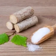 Em uma mesa, há três madeiras empilhadas com duas folhas verdes à frente. Ao lado, temos o xilitol, substituto do açúcar. 