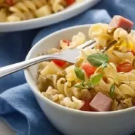 Imagem de um bowl branco com salada de macarrão parafuso com cubos de presunto, tomate e azeitonas.