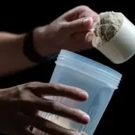 Mulher segurando um copo de plástico com Whey Protein, e com a outra mão segura um medidor cheio da mesma proteína.