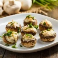 Cogumelos recheados com queijo e ervas frescas servidos em prato branco sobre mesa rústica de madeira.