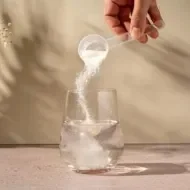 Mão despejando pó de colágeno de uma colher medidora em um copo de água transparente sobre a mesa.