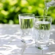 Água sendo servida em copo de vidro sobre mesa branca, com fundo verde desfocado de plantas ao sol.