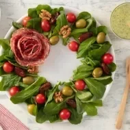 Guirlanda de salada com folhas verdes, tomatinhos, azeitonas, nozes, tâmaras e flor de salame ao centro.