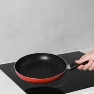 Mão segurando frigideira antiaderente sobre cooktop de indução, pronta para fazer a cura da panela.