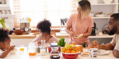 Dicas de café da manhã: Duas crianças negras à esquerda, dois adultos negros à direita, com frutas, leite, pão e café na mesa
