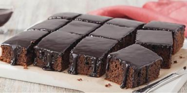 Prato com um bolo de chocolate cortado em pedaços quadrados e uma faca, ao fundo um pano vermelho e um potinho com chocolate 