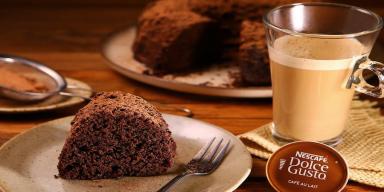 82 receitas doces fáceis para descomplicar a vida Imagem de uma fatia de bolo de chocolate com uma xícara de café com leite e uma cápsula de Dolce Gusto.