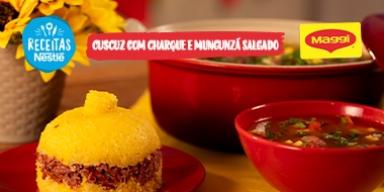 Fotografia mostra um cuscuz recheado com carne seca e um caldo com carnes, ambos em uma mesa de madeira e um pano amarelo.