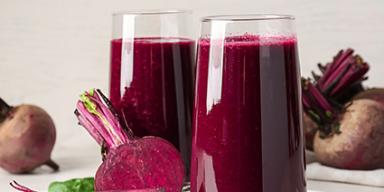 Copos com suco de beterraba, rodeados por beterrabas frescas cortadas e folhas verdes sobre superfície clara.
