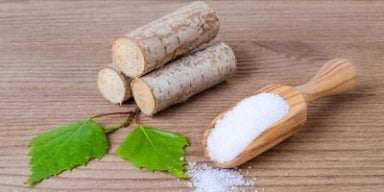 Benefícios do Xilitol Em uma mesa, há três madeiras empilhadas com duas folhas verdes à frente. Ao lado, temos o xilitol, substituto do açúcar.