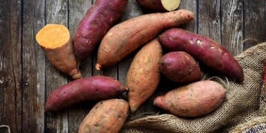 Benefícios da batata-doce para sua saúde Batatas-doces inteiras e cortadas sobre superfície de madeira rústica com saco de estopa ao lado.