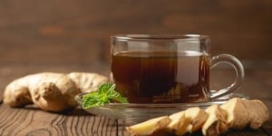 Gengibres inteiros e fatiados ao lado de uma xícara de chá de gengibre. Todos os itens estão em cima de uma mesa de madeira. 