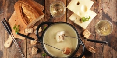 Como fazer fondue Imagem de uma mesa de madeira com uma panela de fondue e acompanhamentos como pães, queijos e vinho.