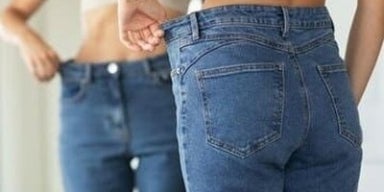 6 razões pelas quais você não consegue emagrecer Mulher se olhando no espelho segurando a sobra da calça jeans na lateral. A imagem representa porque não consigo emagrecer.