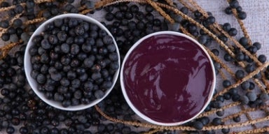 Benefícios do açaí: por que o açaí faz bem? Tigela com polpa de açaí e tigela com frutas de açaí inteiras, sobre ramos com frutos dispostos em superfície clara.