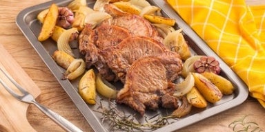 Bisteca: dicas e receitas Imagem de uma assadeira com bistecas, batatas, cebola e alho sobre uma mesa de madeira.