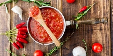 Imagem de uma panela com molho de tomate com uma colher de pau sobre uma superfície de madeira com tomates.
