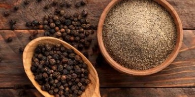 Grãos de pimenta-do-reino em uma colher de madeira e pimenta moída em um pote sobre uma superfície rústica de madeira.