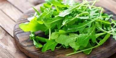 7 benefícios da rúcula Folhas frescas de rúcula com gotas de água sobre uma tábua de madeira rústica.