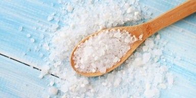 Sal marinho: existem benefícios na alimentação? Colher de madeira com sal matinho sobre superfície azul, com grãos espalhados ao redor.