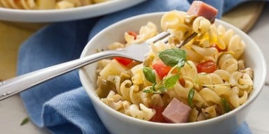 Imagem de um bowl branco com salada de macarrão parafuso com cubos de presunto, tomate e azeitonas.