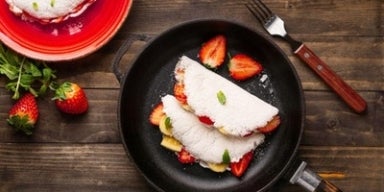 Quais são os benefícios da tapioca? Duas tapiocas recheadas com frutas em uma frigideira sobre uma mesa de madeira. Ao lado, um prato com outra tapioca.