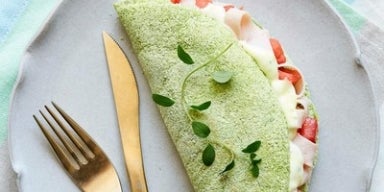 Imagem de uma tapioca verde de espinafre recheada com queijo e tomate sobre um prato cinza.