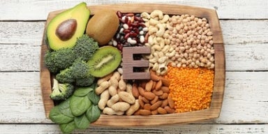 Vitamina E: benefícios e onde encontrar Alimentos ricos em vitamina E em bandeja de madeira: abacate, kiwi, brócolis, espinafre, castanhas e leguminosas.