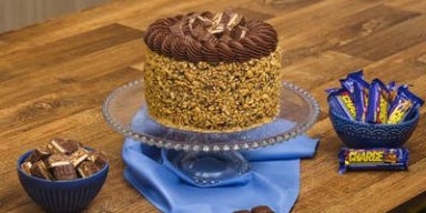 Bolo de chocolate Charge coberto com amendoim triturado e pedaços do chocolate, decorado em prato de vidro.