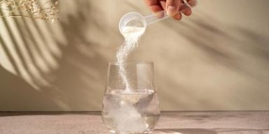 Colágeno: um aliado prático para incluir no dia a dia Mão despejando pó de colágeno de uma colher medidora em um copo de água transparente sobre a mesa.