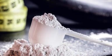 O que é e para que serve a creatina? Colher medidora com creatina em pó branca sobre a mesa, ao lado de uma fita métrica amarela desfocada.