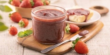 Pote de geleia de morango sobre tábua de madeira, com morangos frescos e pão com geleia ao fundo.