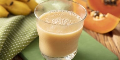 Vitamina cremosa servida em copo transparente, com bananas e fatias de mamão ao fundo sobre mesa de madeira.
