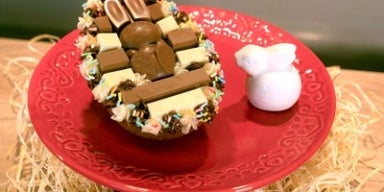Ovo de Páscoa recheado e decorado com chocolates KitKat e confeitos coloridos, servido em prato vermelho com coelho branco.