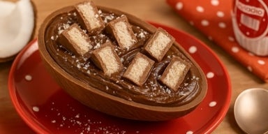 Ovo de chocolate recheado com creme e decorado com pedaços de prestígio, servido em prato vermelho com poás brancos.