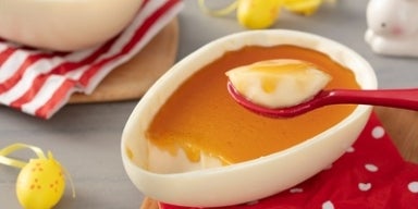 Ovo de Páscoa de chocolate branco recheado com pudim e calda dourada, servido em colher vermelha.