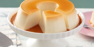 Pudim de leite condensado em forma redonda com calda de caramelo, servido em prato branco sobre mesa clara. 
