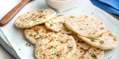 Imagem de pães achatados dourados servidos em prato branco, decorados com salsa e acompanhados de molho em tigela.