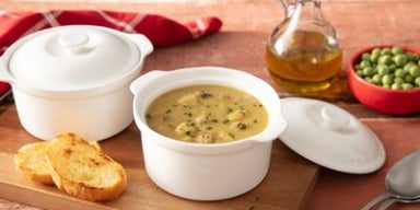 Tigela de sopa de ervilha com pedaços de bacon e cheiro-verde, acompanhada de torradas, azeite e ervilhas frescas ao fundo.
