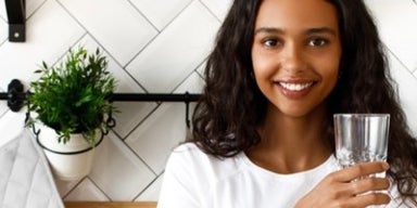 Mulher negra de cabelos longos ondulados está segurando um copo de água para responder a pergunta se suco substitui água.