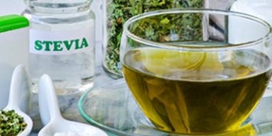 Guia completo sobre os tipos de adoçantes Uma xícara com chá, rodeada por folhas, ervas e especiarias, além de tipos de adoçantes - como a stevia.