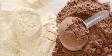 Whey Protein: para que serve e como incluir no dia a dia Colheres medidoras com whey protein em pó sabor baunilha e chocolate, sobre a superfície de pó.