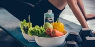 Mulher com roupas de ginástica sentada em um tapete no chão da academia. Ao lado, uma tigela com legumes e Halteres.