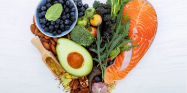 Imagem de alimentos saudáveis dispostos em forma de coração, incluindo salmão, abacate, mirtilos e nozes sobre fundo branco.